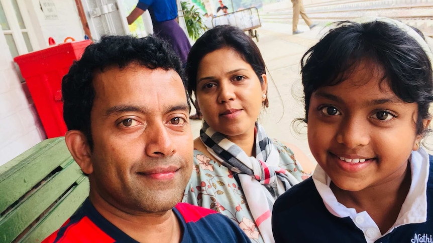 Sudesh Kolonne wears a striped shirt and takes a selfie with his wife and daughter smiling.