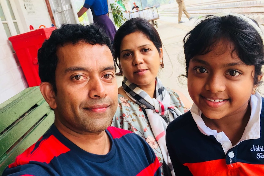 Sudesh Kolonne wears a striped shirt and takes a selfie with his wife and daughter smiling.