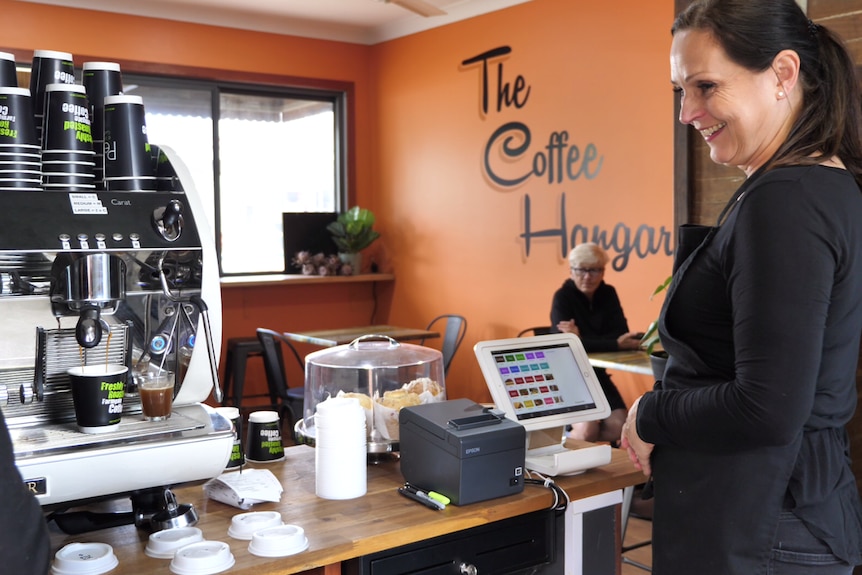 A woman with dark hair stands behind the counter of a cafe, smiling.