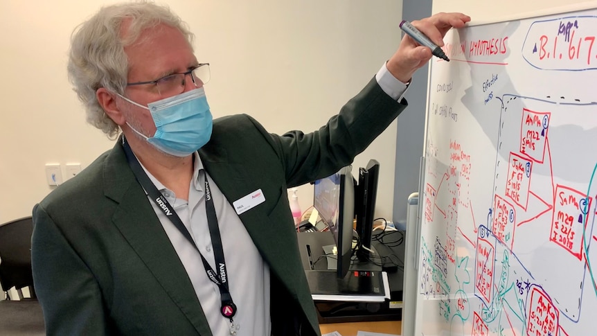 A grey-haired man in a suit jacket and light shirt wears a mask and writes on a whiteboard.