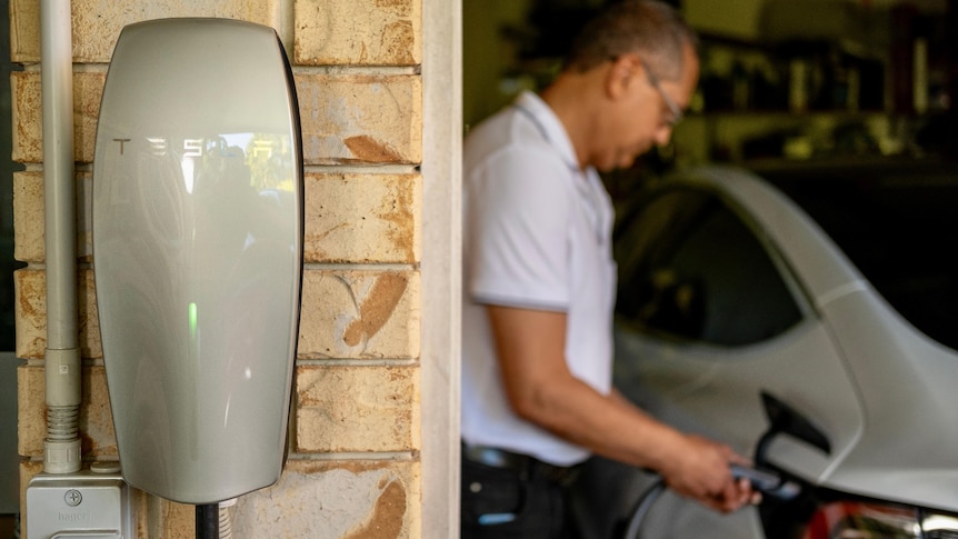 a close-up of an electric vehicle charger mounted to a garage wall. a man is charging his car in the background