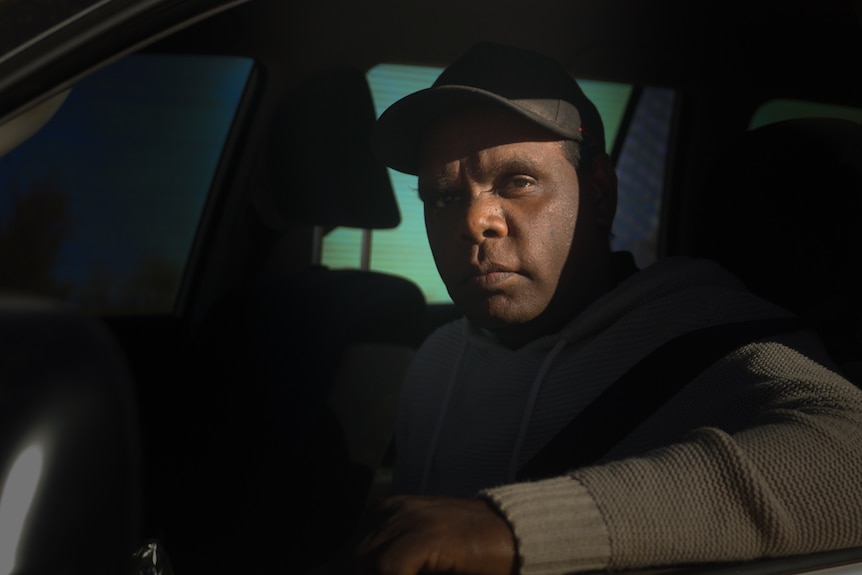 A man looks out from the window of a car, wearing a baseball cap.