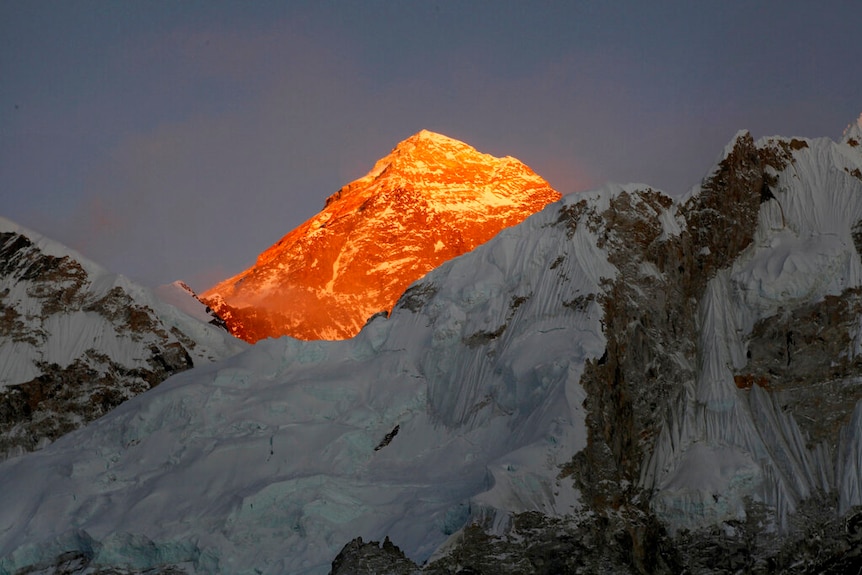 Looking up, you view the peak of Mount Everest bathed in golden sunlight against a dimly lit clear sky.