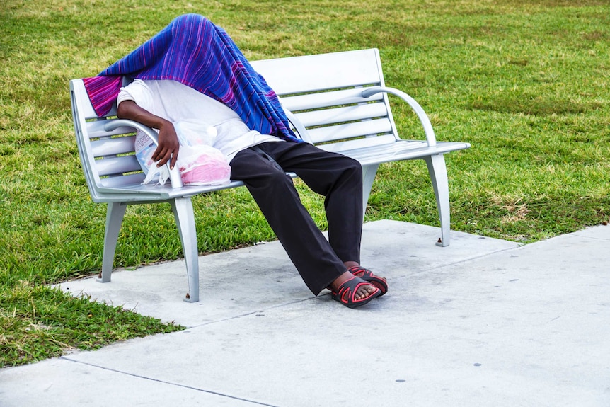 A homeless person on bench with a arm rest in the middle, to prevent people lying down.