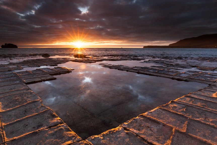 The sun sets over water and square shaped rocks on the coast of Tasmania.
