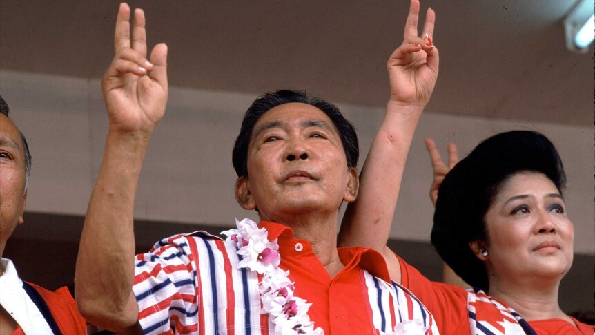 Marcos and his wife, both wearing bright red, look out on a crowd holding their right hands up in the peace sign.