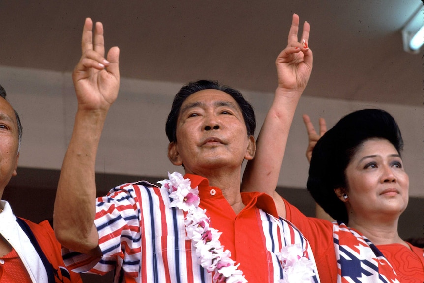 Marcos and his wife, both wearing bright red, look out on a crowd holding their right hands up in the peace sign.