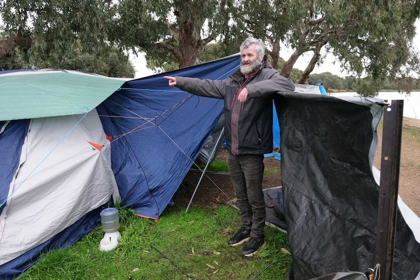 A man with grey hair and a beard, wearing black jeans and a black jacket, stands in front of a tent pointing. 
