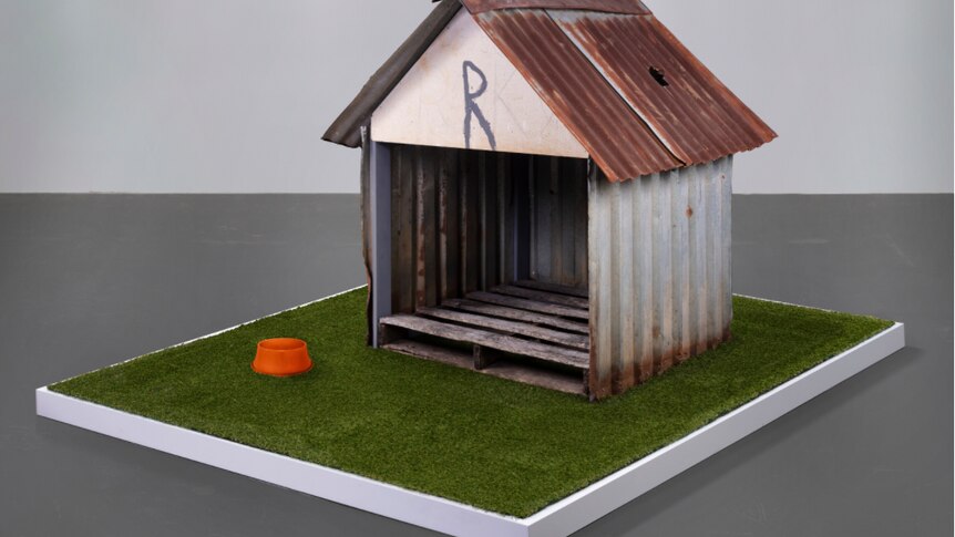 A damaged modest doghouse sits atop a astroturf-covered display base in a large plain room.