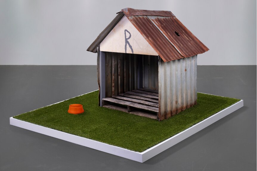 A damaged modest doghouse sits atop a astroturf-covered display base in a large plain room.