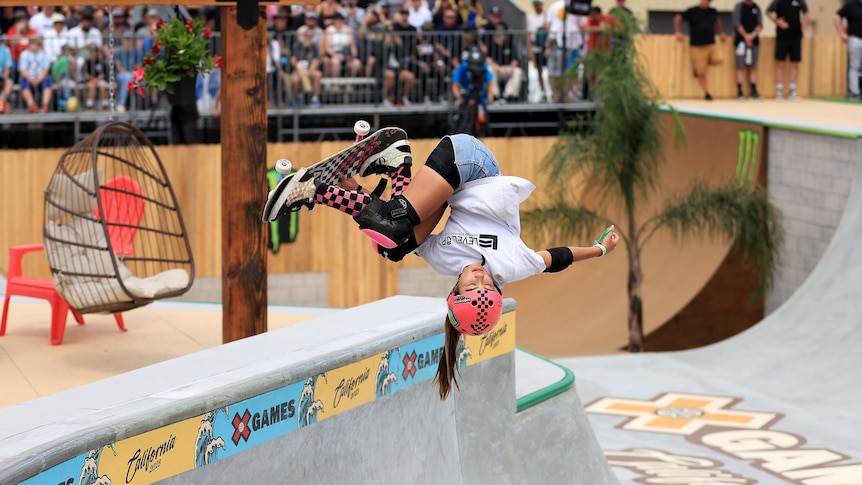 Skateboarder Arisa Trew is upside down, holding her skateboard as she performs a trick.