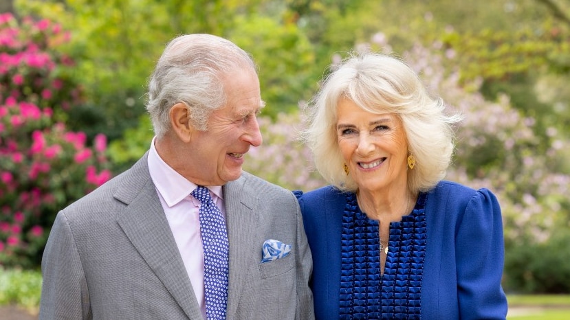 A man in a suit standing outside links arms with a woman and gazes at her, while she smiles at the camera