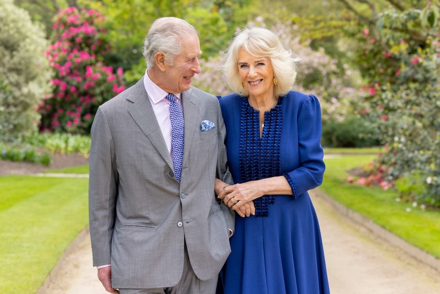 A man in a suit standing outside links arms with a woman and gazes at her, while she smiles at the camera