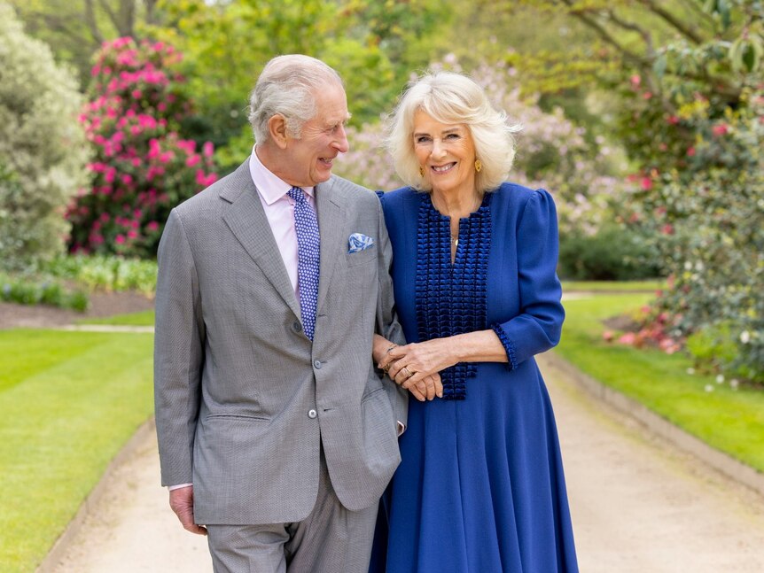 A man in a suit standing outside links arms with a woman and gazes at her, while she smiles at the camera