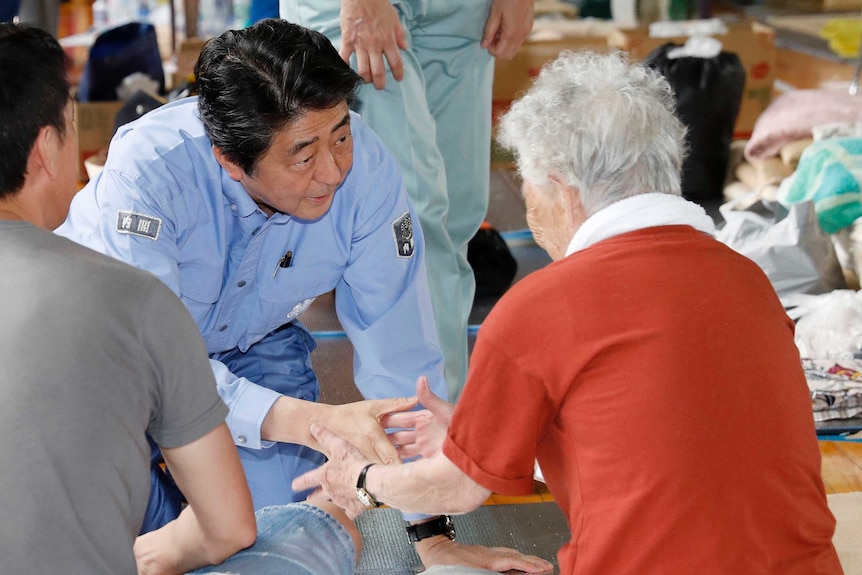 Shinzo Abe sits on his knees as he shakes hands and talks to an elderly woman.