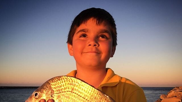A young boy holds a fish he caught, with the ocean and sunset in the background.
