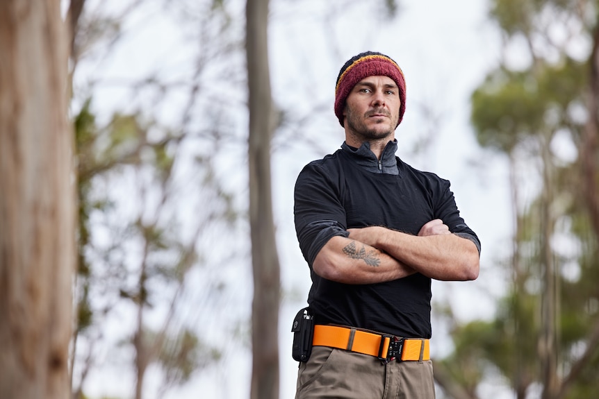 A man in his mid 30s in a beanie with the colours of the Aboriginal flag in a dark top, arms crossed, orange belt, forest behind