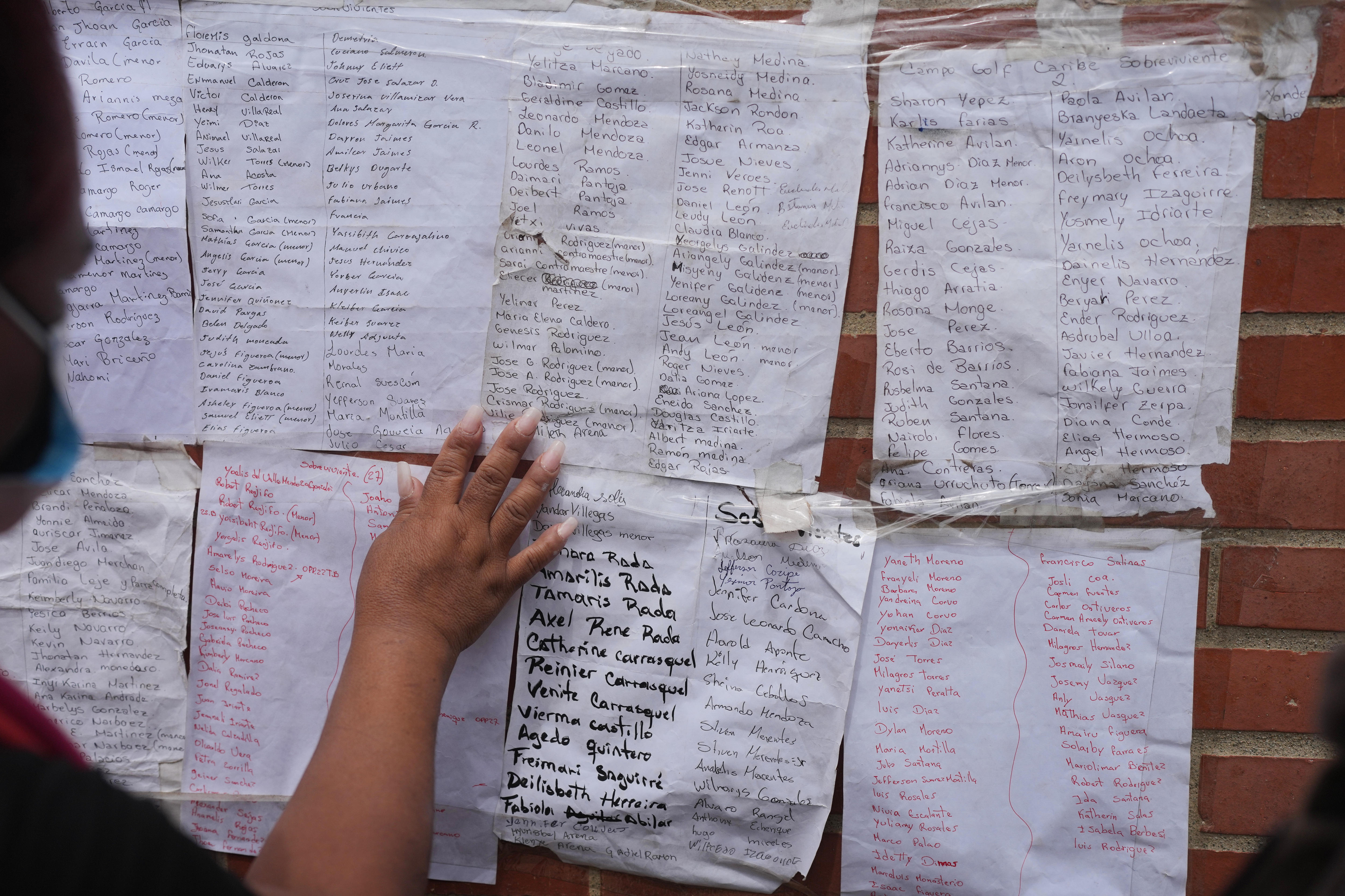 A blurred woman's silhouette, while she place her hands on white paper lists of names on a brick wall.