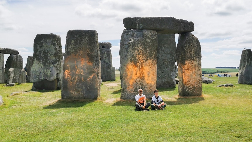Two people sit on ground in front of orange stonehenge 