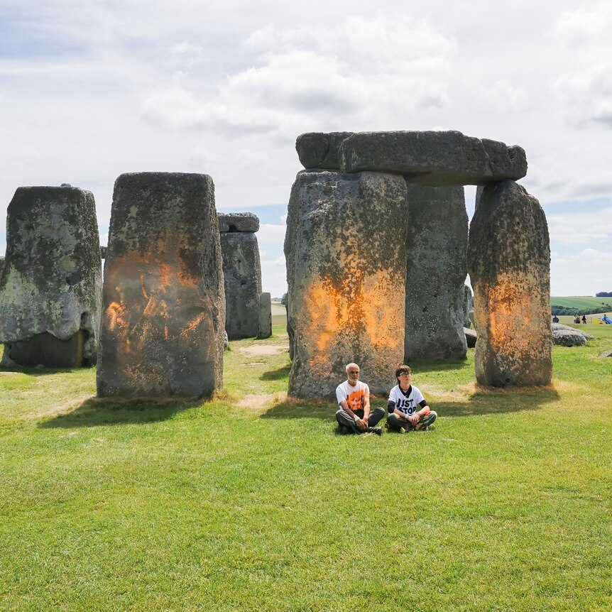 Two people sit on ground in front of orange stonehenge 