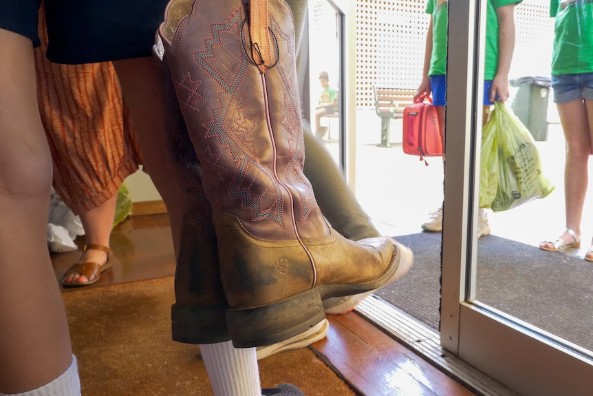 High top boots held in the foreground as kids line up outside a glass doors, Longreach, November 2022.