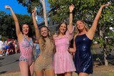 A group of four girls smile and pose with one hand each in the air. They're all wearing glittery bodysuits or dresses