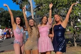A group of four girls smile and pose with one hand each in the air. They're all wearing glittery bodysuits or dresses