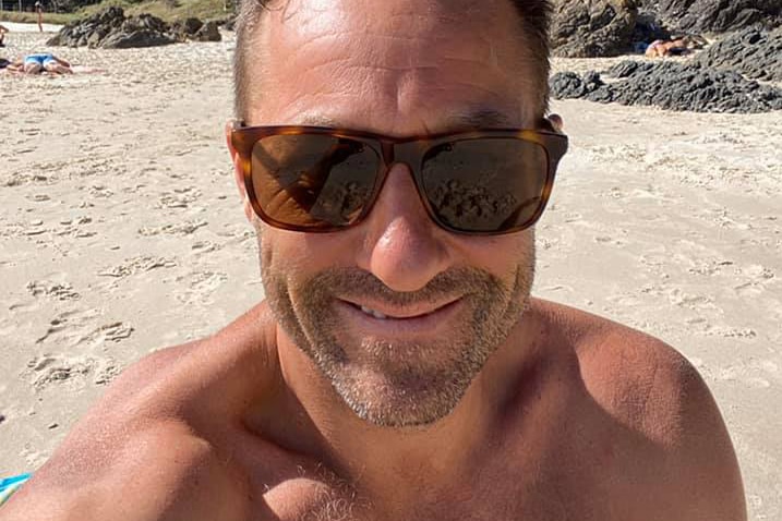 A tight head and shoulders shot of a man at the beach wearing sunglasses and smiling.