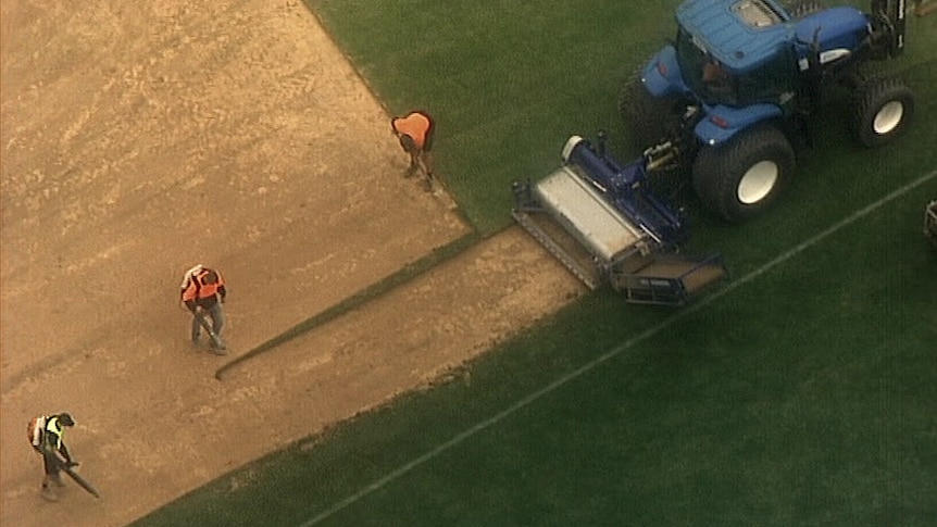 Three men work on a dirst and grassed playing field next to machinery.