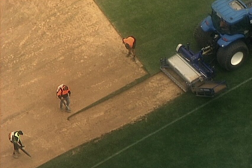 Three men work on a dirst and grassed playing field next to machinery.
