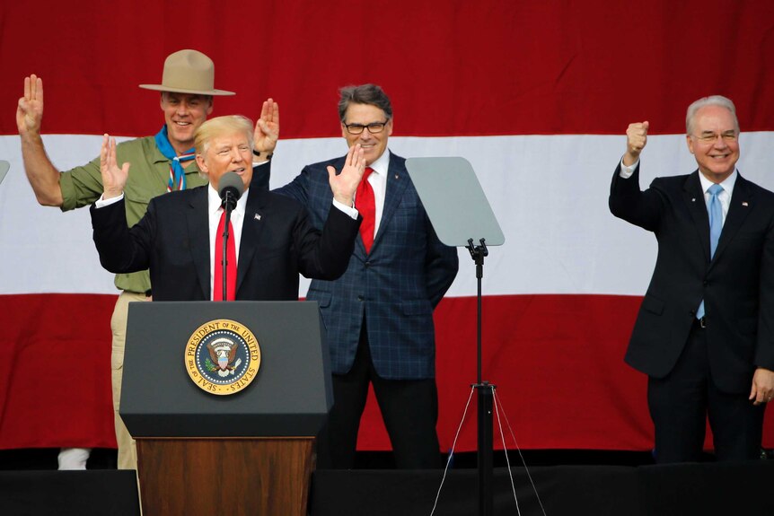 US president gestures behind a microphone. He is flanked by a man dressed in a scouts uniform and two other men.