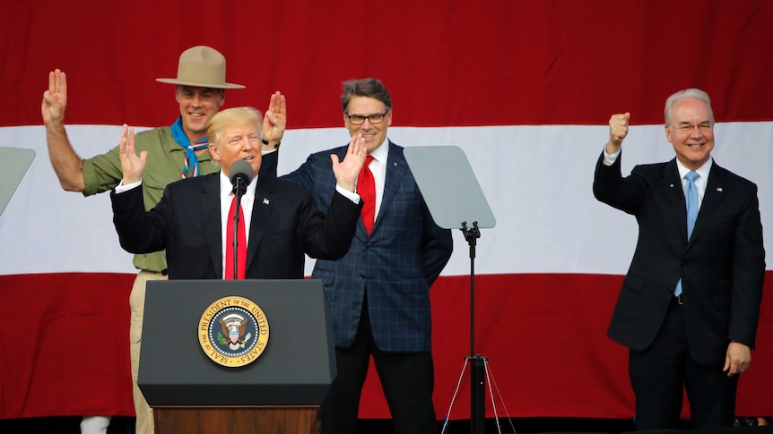 US president gestures behind a microphone. He is flanked by a man dressed in a scouts uniform and two other men.