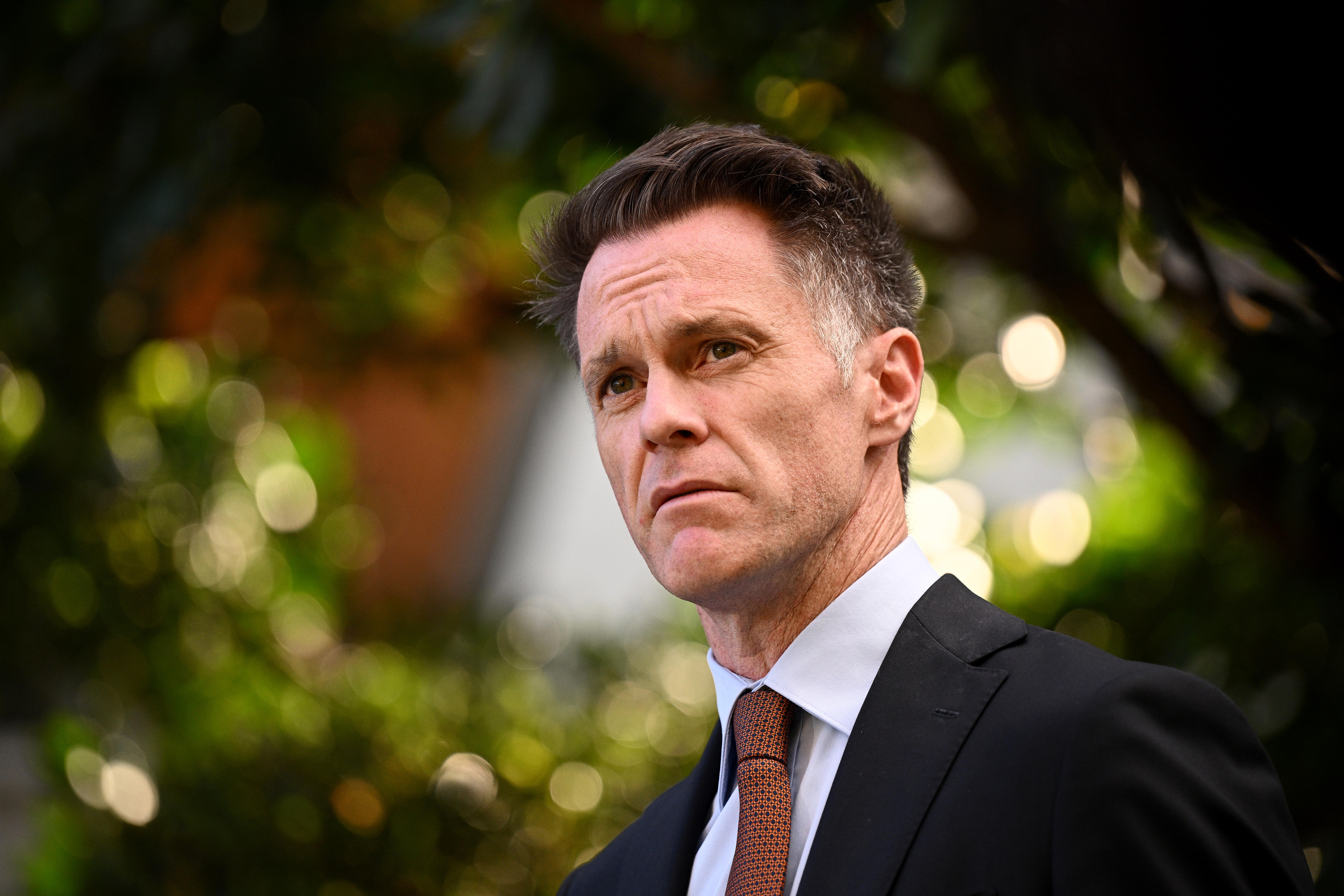 A serious dark-haired man in a dark suit – Chris Minns – stands near some trees, background is blurred.