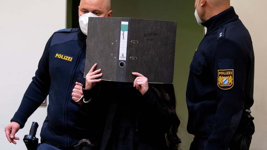 Woman in black escorted by two police in face masks covers face with black folder