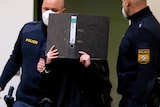 Woman in black escorted by two police in face masks covers face with black folder