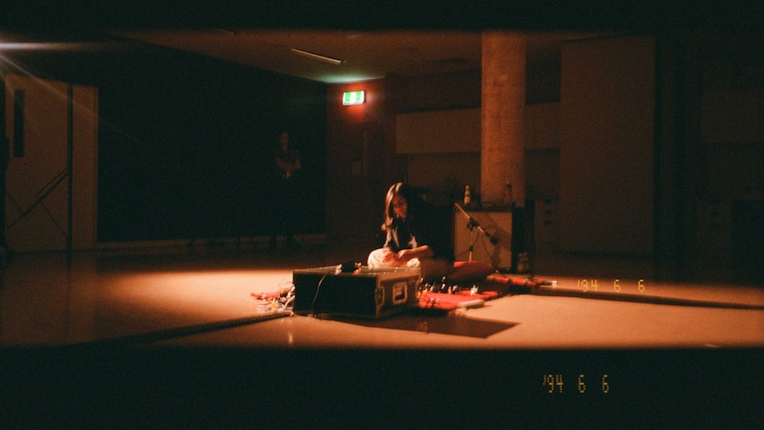 A film photo of Sia sitting on the floor of a dark room with cables and and amplifier in front of her.
