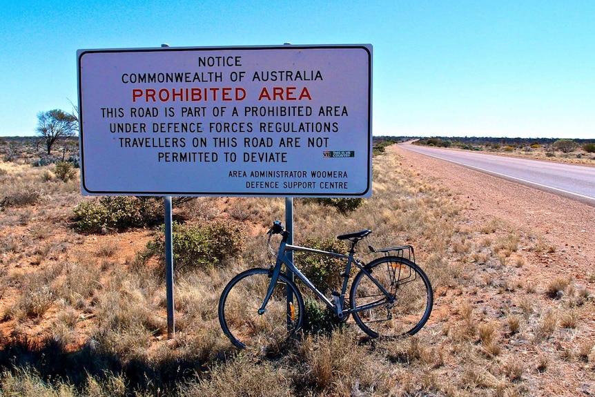 A white sign that says 'prohibited area' in the middle of the outback.  There is a bike in front of it.