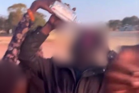A person, face blurred, drinking from a glass bottle with another person's hand tipping the bottle