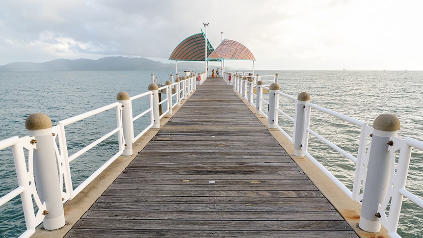 A timber jetty extends out into the ocean, colourful shade sails provide shade in the distance.