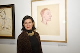 Paloma Picasso, with shoulder-length brown hair and brown jacket, smiles with closed mouth near large drawing in frame.