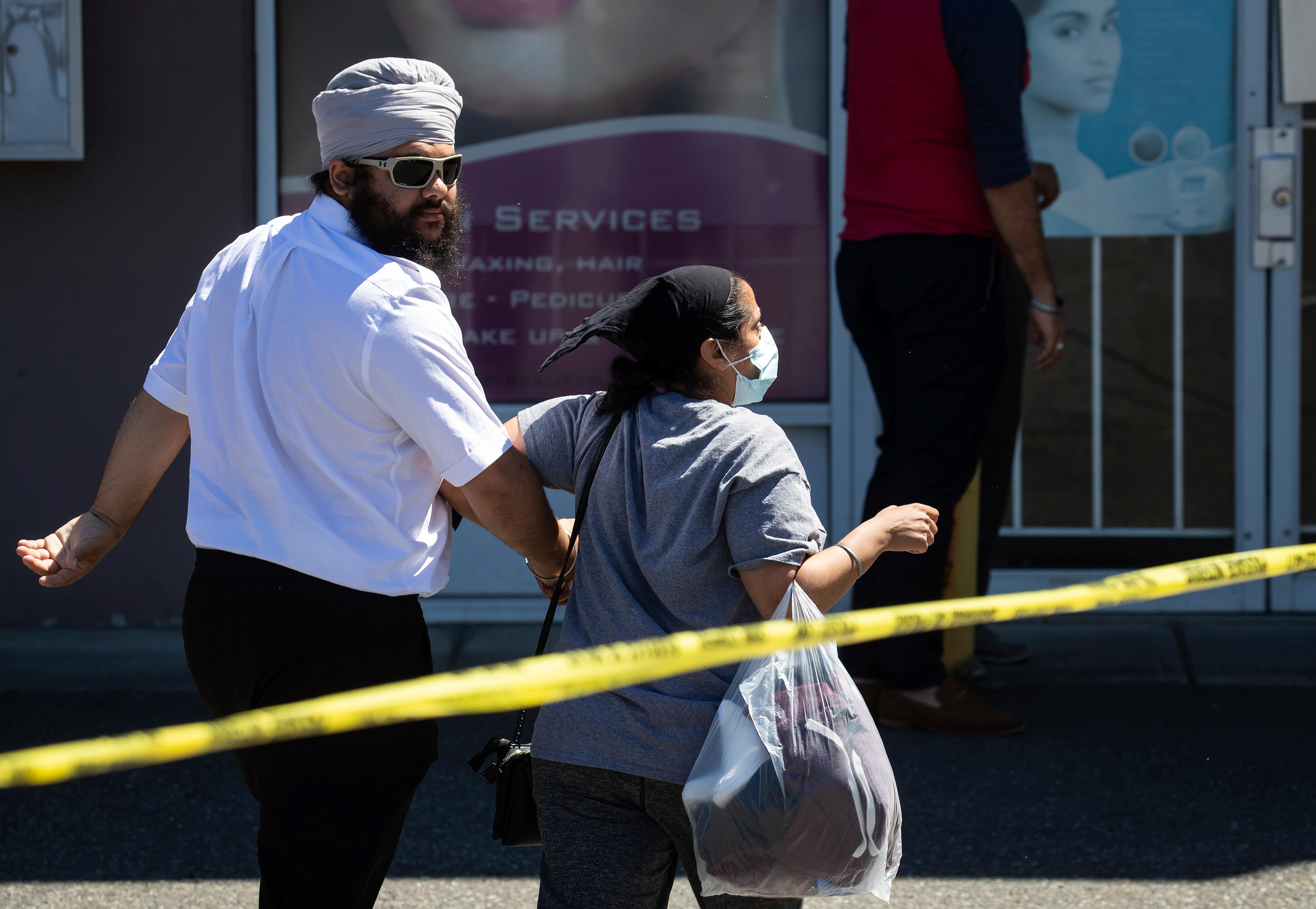 A man and a woman walk past yellow police tape. The man looks over his shoulder at the camera with a blank expression. 