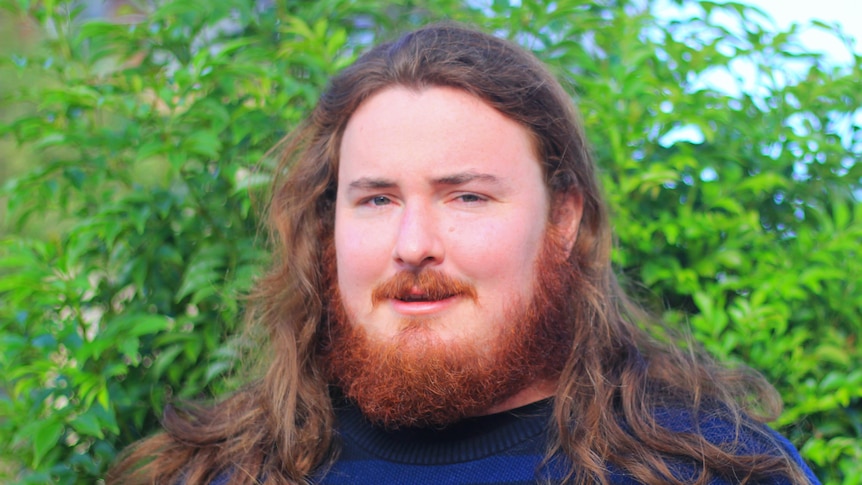 A man with a red beard and hair, in front of a green tree, with a blue jumper on. He is serious.