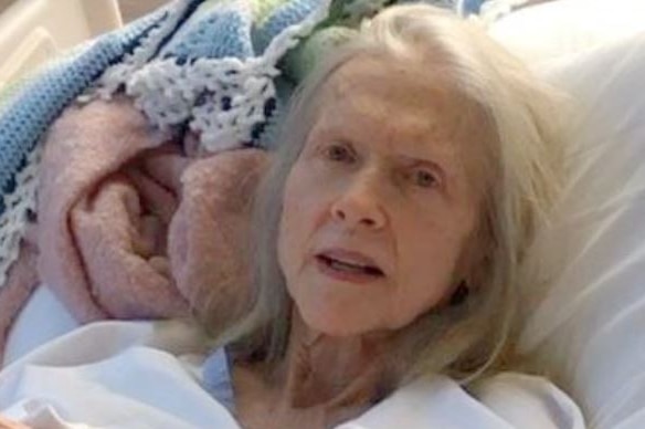 An elderly woman with long white hair lying in a hospital bed.