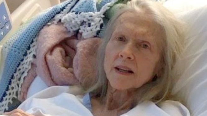 An elderly woman with long white hair lying in a hospital bed.