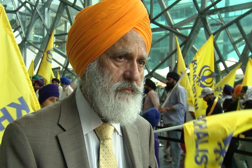 A Sikh man standing in front of a protest with yellow and blue flags