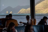 People sitting on a boat look out at a mountain and some rocks protruding from the ocean.