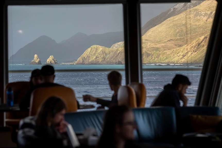 People sitting on a boat look out at a mountain and some rocks protruding from the ocean.