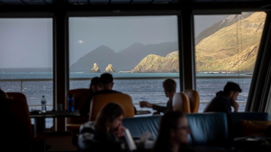 People sitting on a boat look out at a mountain and some rocks protruding from the ocean.