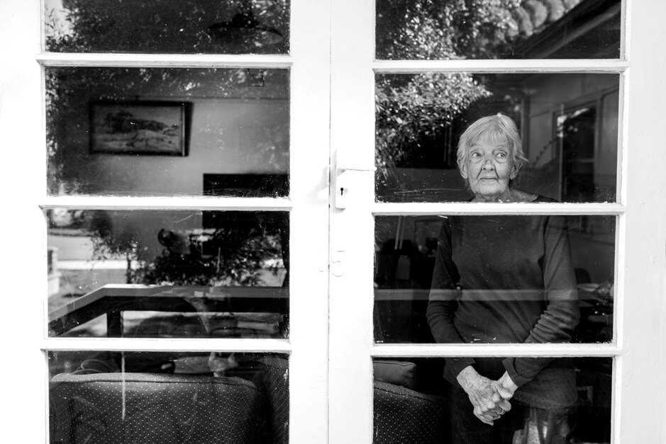 Black and white photo of elderly woman inside a house looking outside through a glass door.