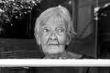 Black and white photo of elderly woman inside a house looking outside through a glass door.